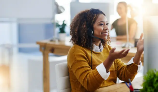 A woman in a brown sweater engages in a discussion at a modern workspace, wearing a headset and gesturing with her hands.