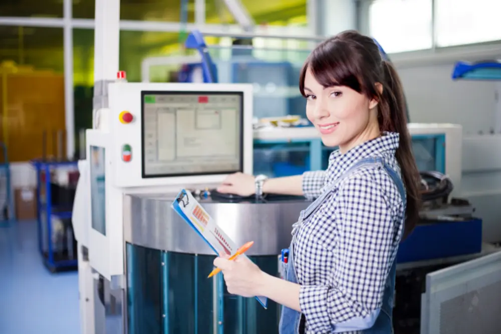 A woman stands by a machine with a computer screen and clipboard, monitoring equipment in a factory setting.