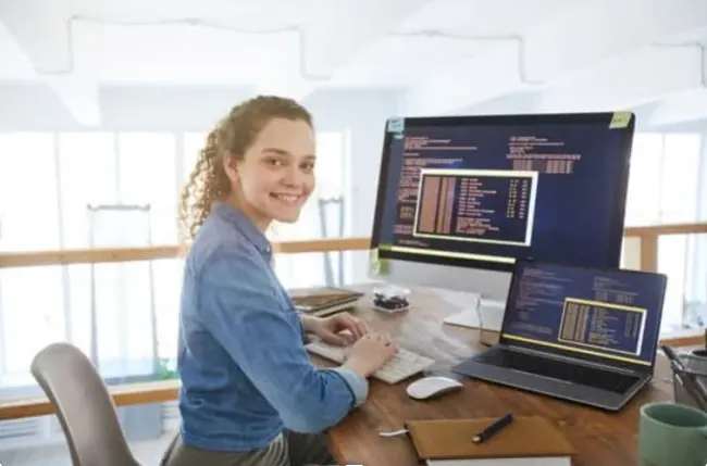 A person with curly hair works at a wooden desk, focused on coding displayed on two computer monitors in a bright, modern workspace