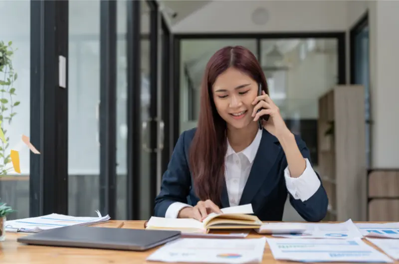 A woman in a business suit sits at a desk, talking on the phone while reviewing notes and documents in a modern office setting.