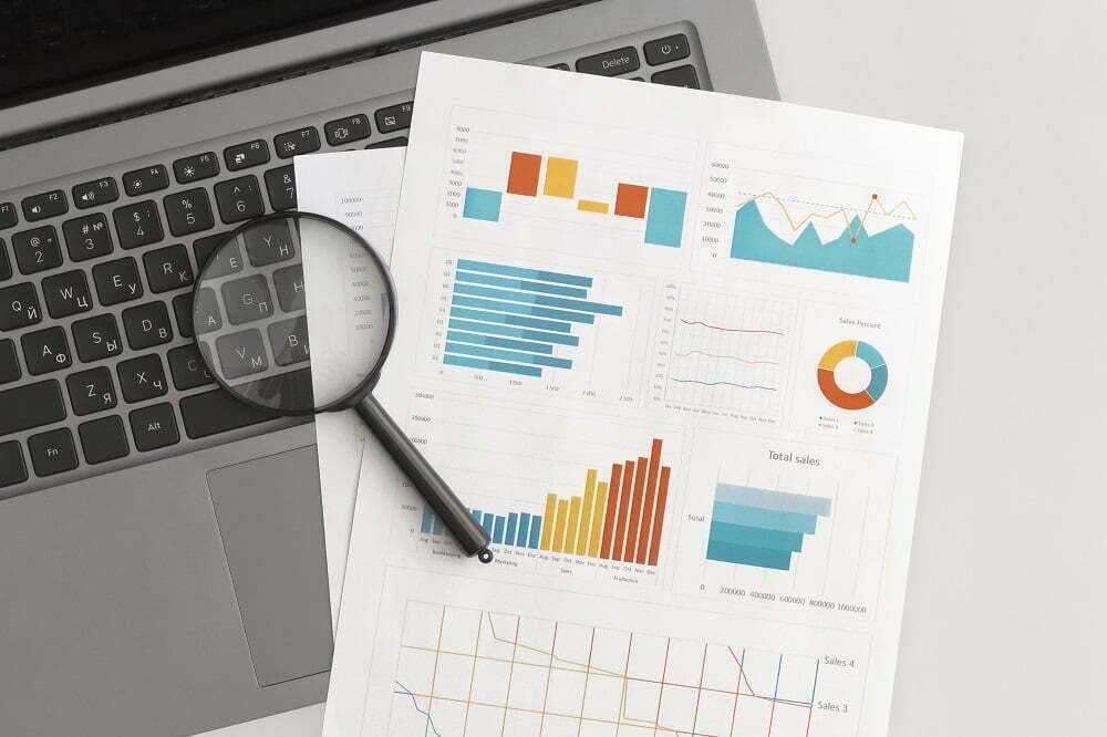Close-up of a laptop keyboard with printed sales graphs and charts, alongside a magnifying glass for analysis.