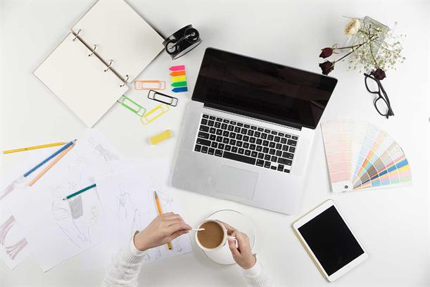 A workspace with a laptop, coffee cup, sketches, stationery, color swatches, tablet, and a notebook.