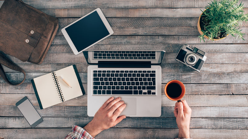 Overhead view of a workspace: a laptop, tablet, notebook, smartphone, camera, coffee cup, and a potted plant on a wooden table.