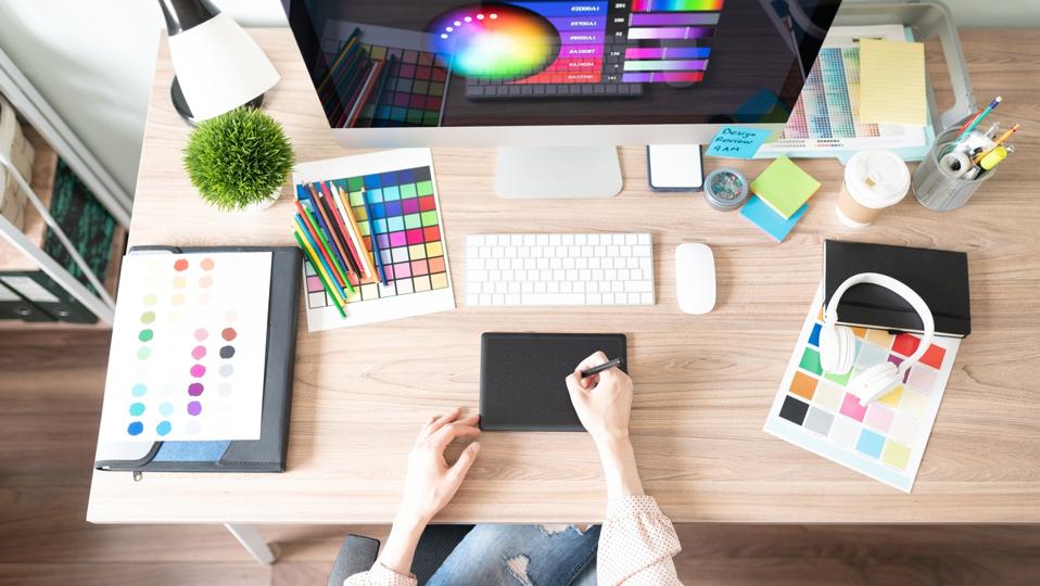 A creative workspace featuring a person drawing on a tablet, colorful palettes, a computer, and art supplies on a wooden desk.