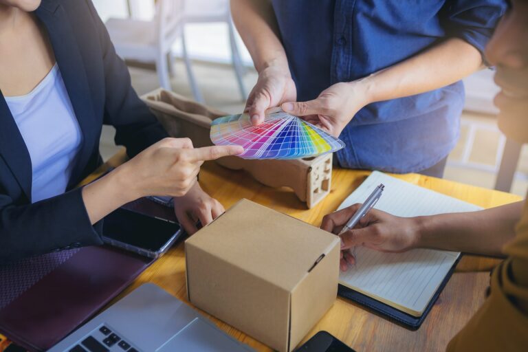 Three designers collaborate at a table, discussing color options from a palette while examining a cardboard box and taking notes.