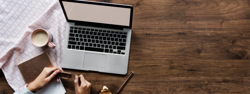 A top-down view of a laptop on a wooden table alongside a coffee cup, notebook, and a piece of chocolate, creating a cozy workspace.