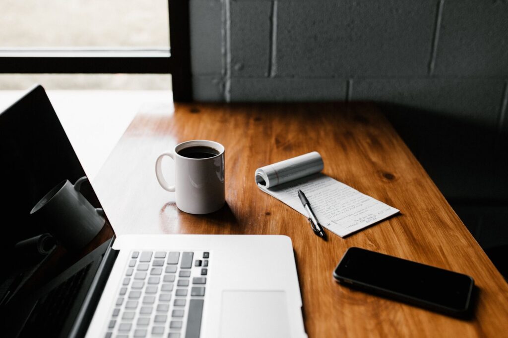 A wooden table features a laptop, a white coffee mug, a notepad with notes, and a smartphone, set near a window.