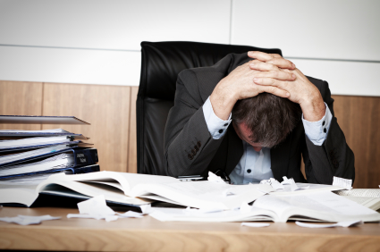 A stressed businessman sitting at a cluttered desk, head in hands, surrounded by piles of papers and documents.