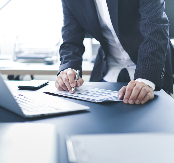 A business professional in a suit analyzes charts on paper while taking notes, with a laptop and phone visible on the desk.