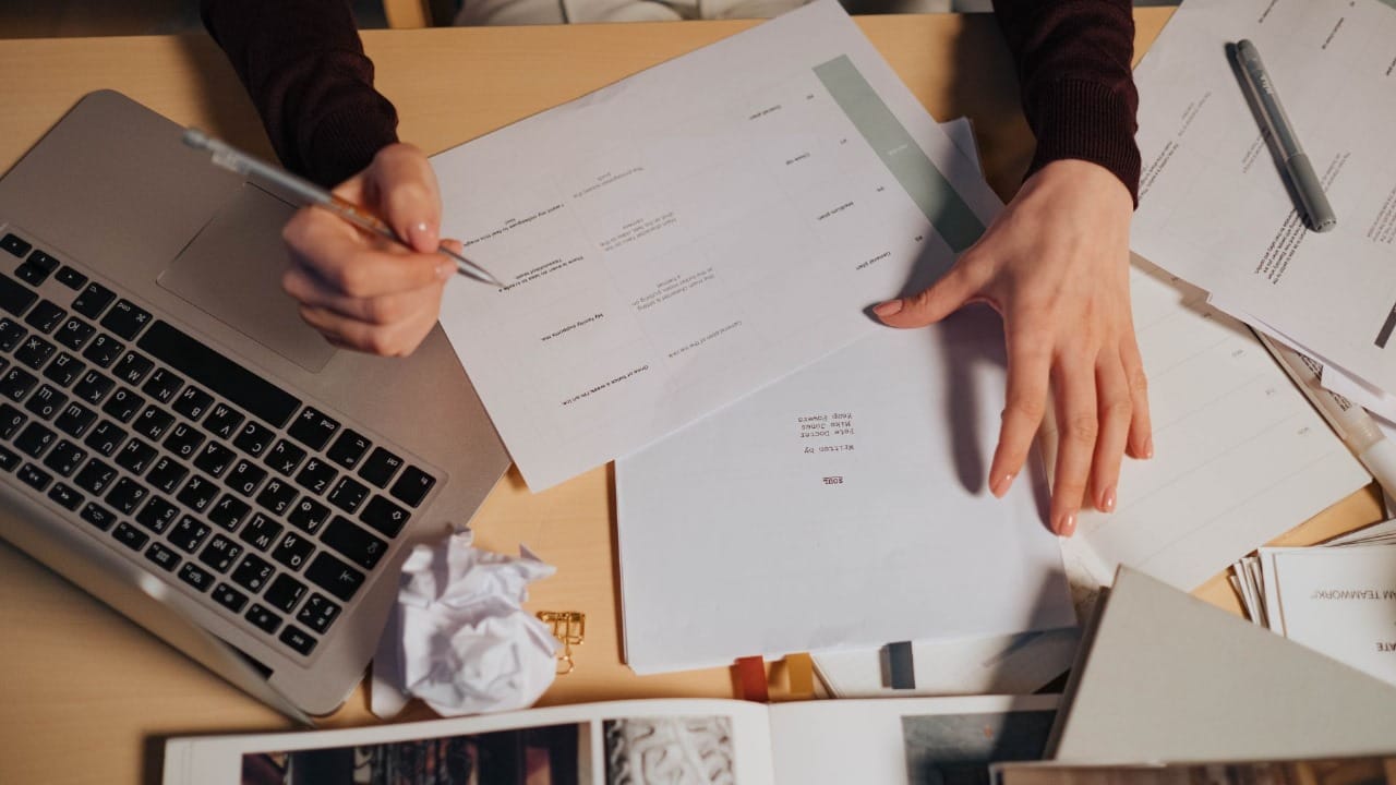 A person reviews printed documents while writing notes, with a laptop and scattered papers on a wooden desk.
