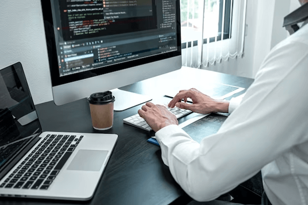 A person in a white shirt typing on a keyboard in front of two computers, one displaying code and a coffee cup nearby.