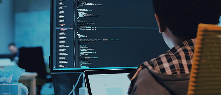 A person in a plaid shirt focused on coding, with lines of programming displayed on a large monitor and a laptop beside them.