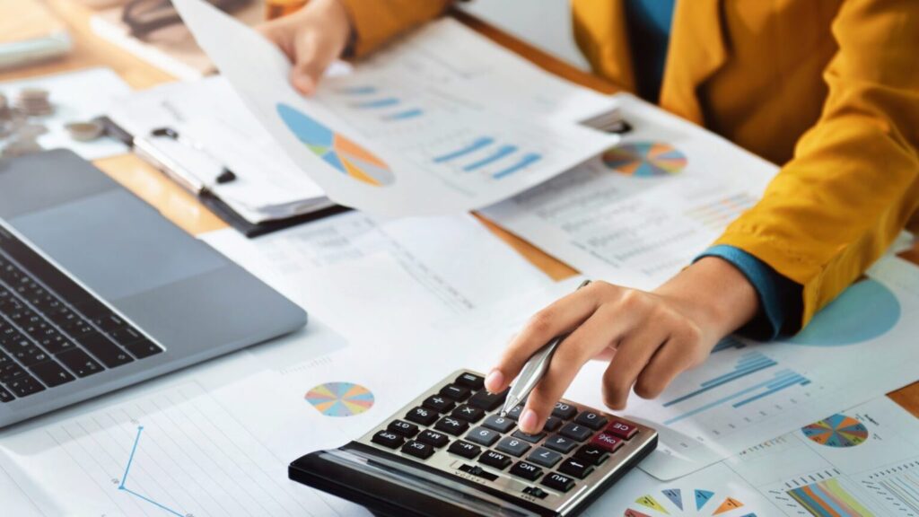 A hand using a calculator amidst a cluttered workspace filled with financial documents and a laptop