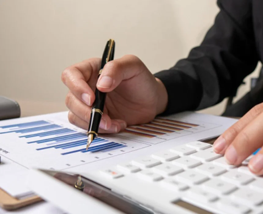 A hand holds a pen over a chart, while the other hand uses a calculator, indicating data analysis or financial calculation.