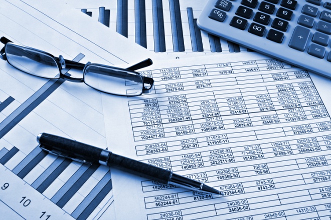 A calculator, pen, and reading glasses are placed on financial spreadsheets with columns of numbers and bar graphs in a blue-toned setting.