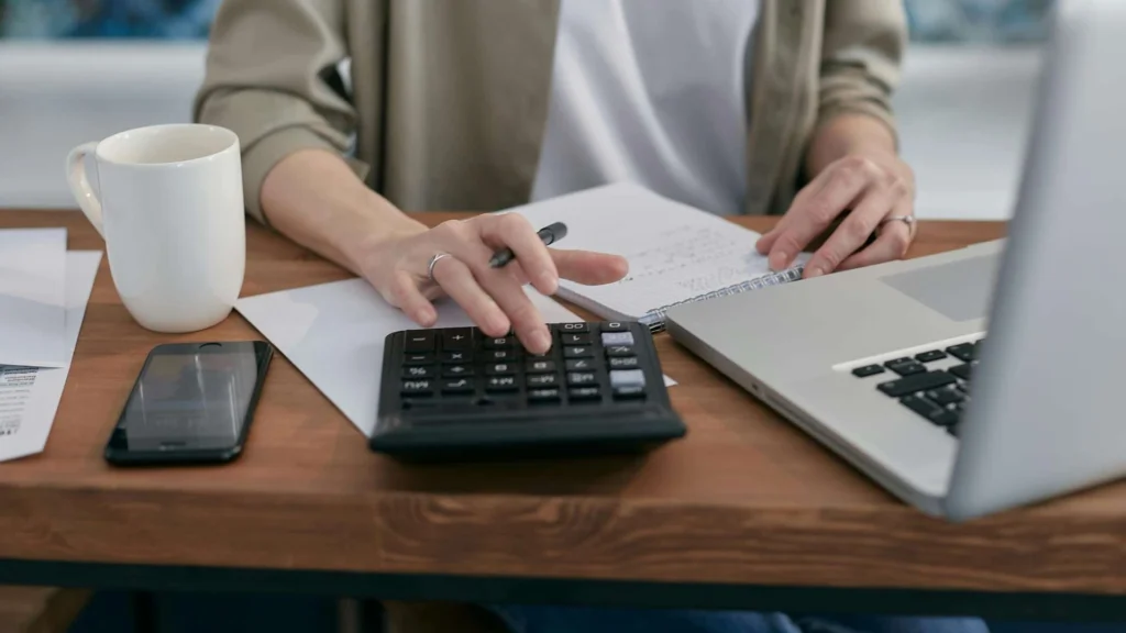 A person calculates with a calculator while sipping coffee, surrounded by papers, a notebook, and a laptop on a wooden desk.