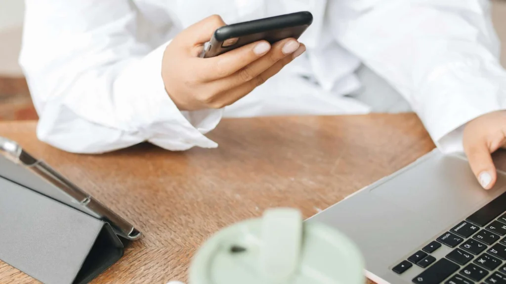 A person in a white shirt holds a smartphone in one hand, while typing on a laptop, with a green cup nearby on a wooden desk.