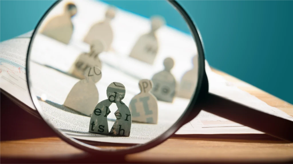 A magnifying glass focuses on wooden letter figures arranged on an open book, symbolizing research and analysis.